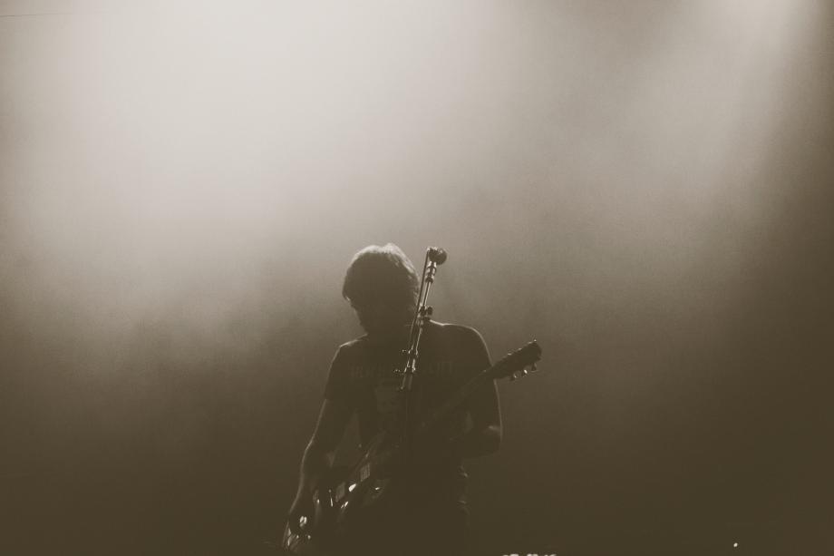 Rockmusik Die Silhouette eines Musikers mit Gitarre, vor ihm steht ein Mikrofonständer, das Bild ist dunkel gehalten.