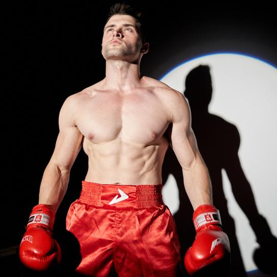 Ein Boxer mir roten Hosen und roten Handschuhen wird von einem Scheinwerfer angestrahlt.