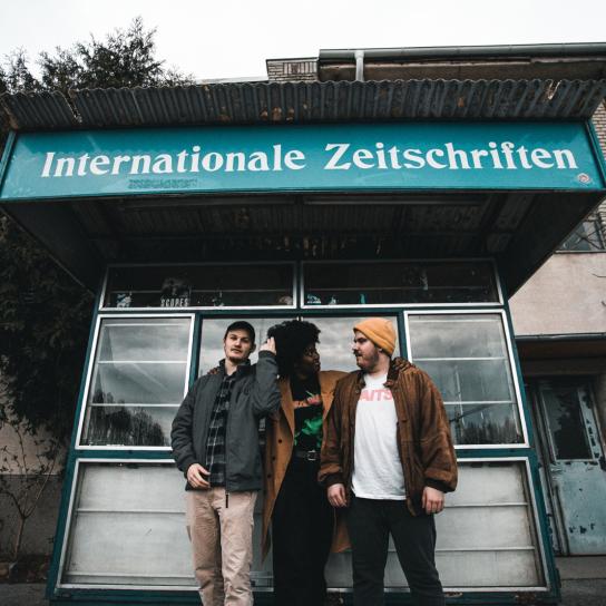 Die drei Mitglieder von Curb stehen vor einem Zeitungsstand mit der Aufschrift "Internationale Zeitschriften""