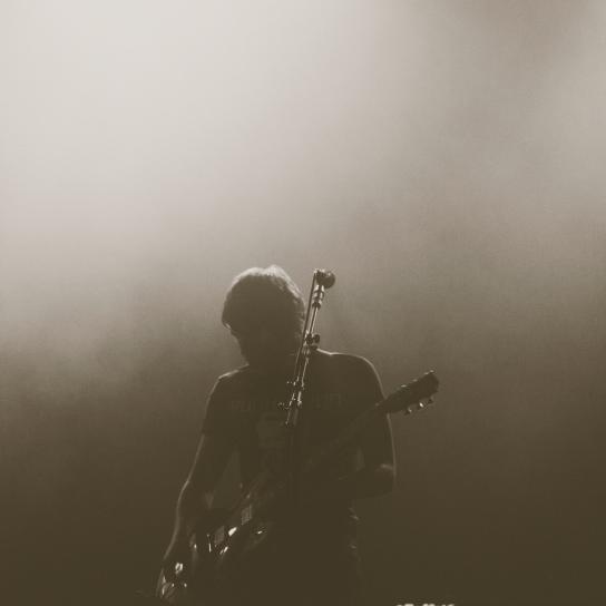 Die Silhouette eines Musikers mit Gitarre, vor ihm steht ein Mikrofonständer, das Bild ist dunkel gehalten.
