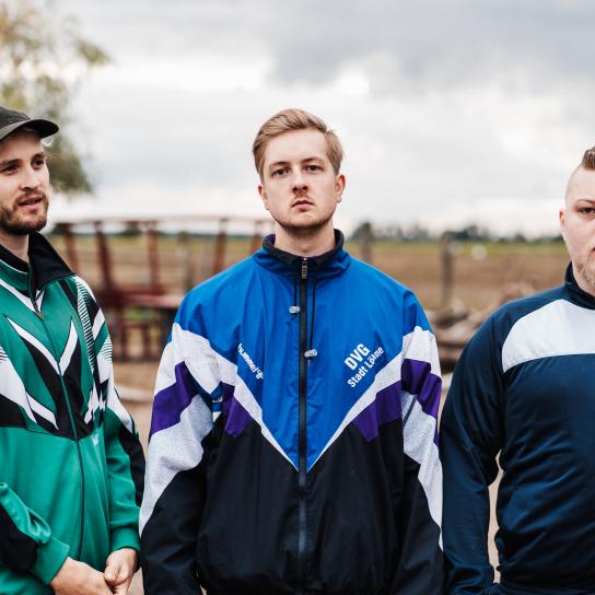 Auf dem Bild stehen die drei Mitglieder der Band Assumption vor einem unscharfen Naturhintergrund.