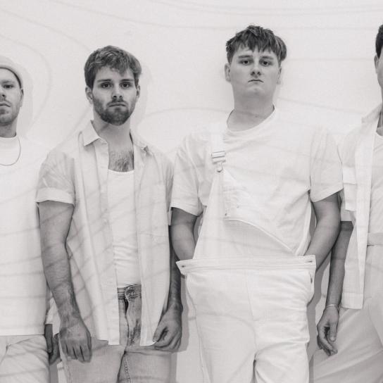 Ein Schwarz Weiß Bild der vierköpfigen Band Razz. Sie stehen nebeneinander an einer Wand und schauen direkt in die Kamera. Alle tragen weiße Kleidung