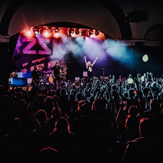 Ein großes Publikum wird nur durch ein paar Scheinwerfer angestrahlt. Das Publkum schauf auf eine Bühne. Auf der Bühne steht Sänger Josh und hebt die Arme in die Luft.