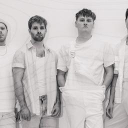 Ein Schwarz Weiß Bild der vierköpfigen Band Razz. Sie stehen nebeneinander an einer Wand und schauen direkt in die Kamera. Alle tragen weiße Kleidung